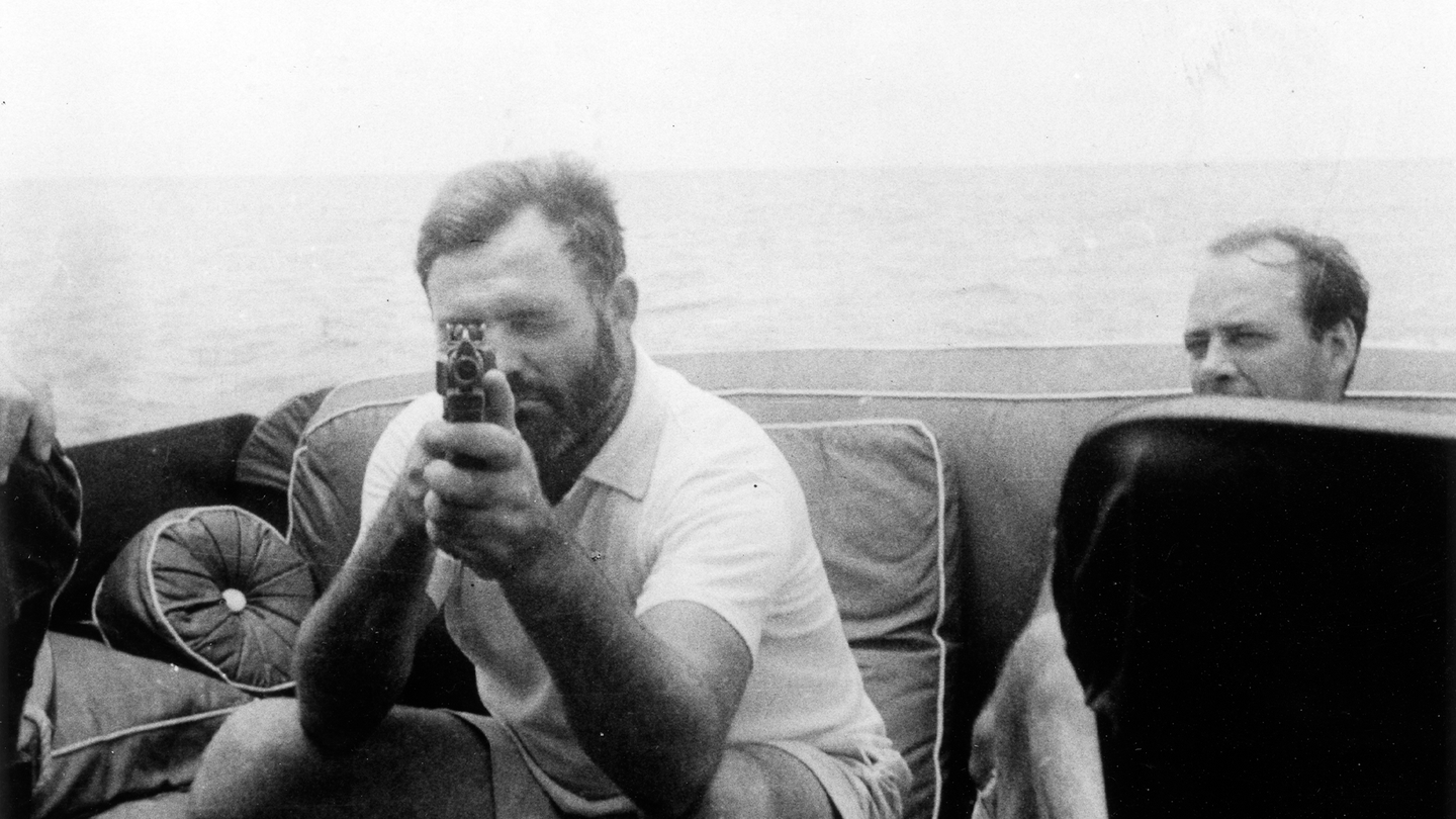 Photograph of Ernest Hemingway, holding a tommy gun, aboard his yacht, the Pilar. Public domain.