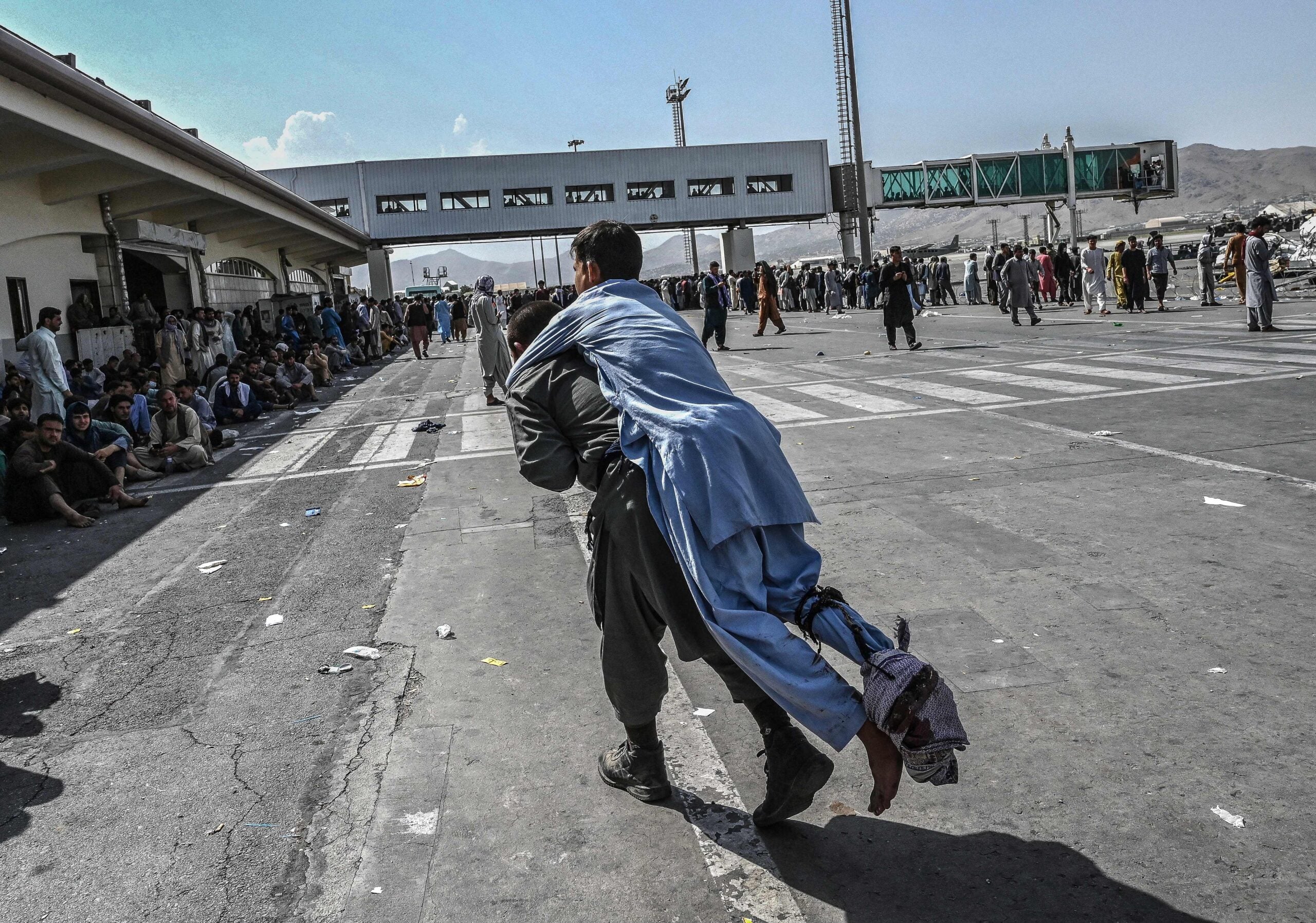 TOPSHOT - A volunteer carries an injured man as other people can be seen waiting at the Kabul airport in Kabul on August 16, 2021, after a stunningly swift end to Afghanistan's 20-year war, as thousands of people mobbed the city's airport trying to flee the group's feared hardline brand of Islamist rule. (Photo by Wakil Kohsar / AFP) (Photo by WAKIL KOHSAR/AFP via Getty Images)