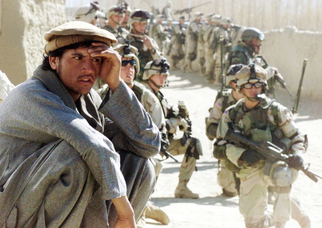 YAYEH KEHL, AFGHANISTAN - NOVEMBER 14:  An Afghani squats on a wall while a group of U.S. Army soldiers from the 82nd Airborne Division secure an area during the inspection of a local bazaar November 14, 2002 in the town of Yayeh Kehl, Paktia province, south of Kabul, Afghanistan. U.S. troops conducted a four-day operation searching for weapons and suspected militant linked to al-Qaeda in the area.  (Photo by Amel Emric-Pool/Getty Images)