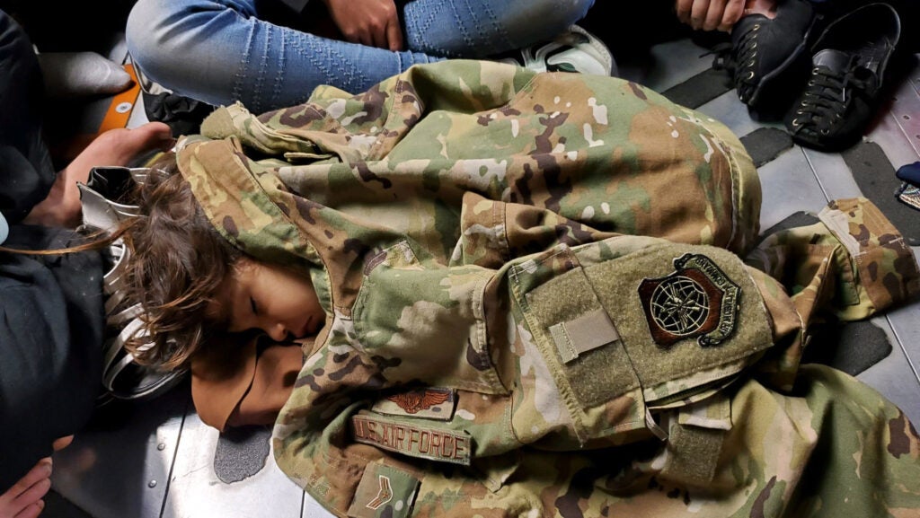 An Afghan child sleeps on the cargo floor of a U.S. Air Force C-17 Globemaster III, kept warm by the uniform of Airman First Class Nicolas Baron, C-17 loadmaster, during an evacuation flight from Kabul, Afghanistan, Aug. 18, 2021. Operating a fleet of Air National Guard, Air Force Reserve and Active Duty C-17s, Air Mobility Command, in support of the Department of Defense, moved forces into theater to facilitate the safe departure and relocation of U.S. citizens, Special Immigration Visa recipients, and vulnerable Afghan populations from Afghanistan. (Photo courtesy 1st Lt. Mark Lawson, 6th Airlift Squadron)