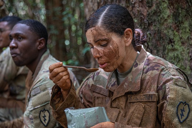 A soldier eating an MRE with a mud covered face.