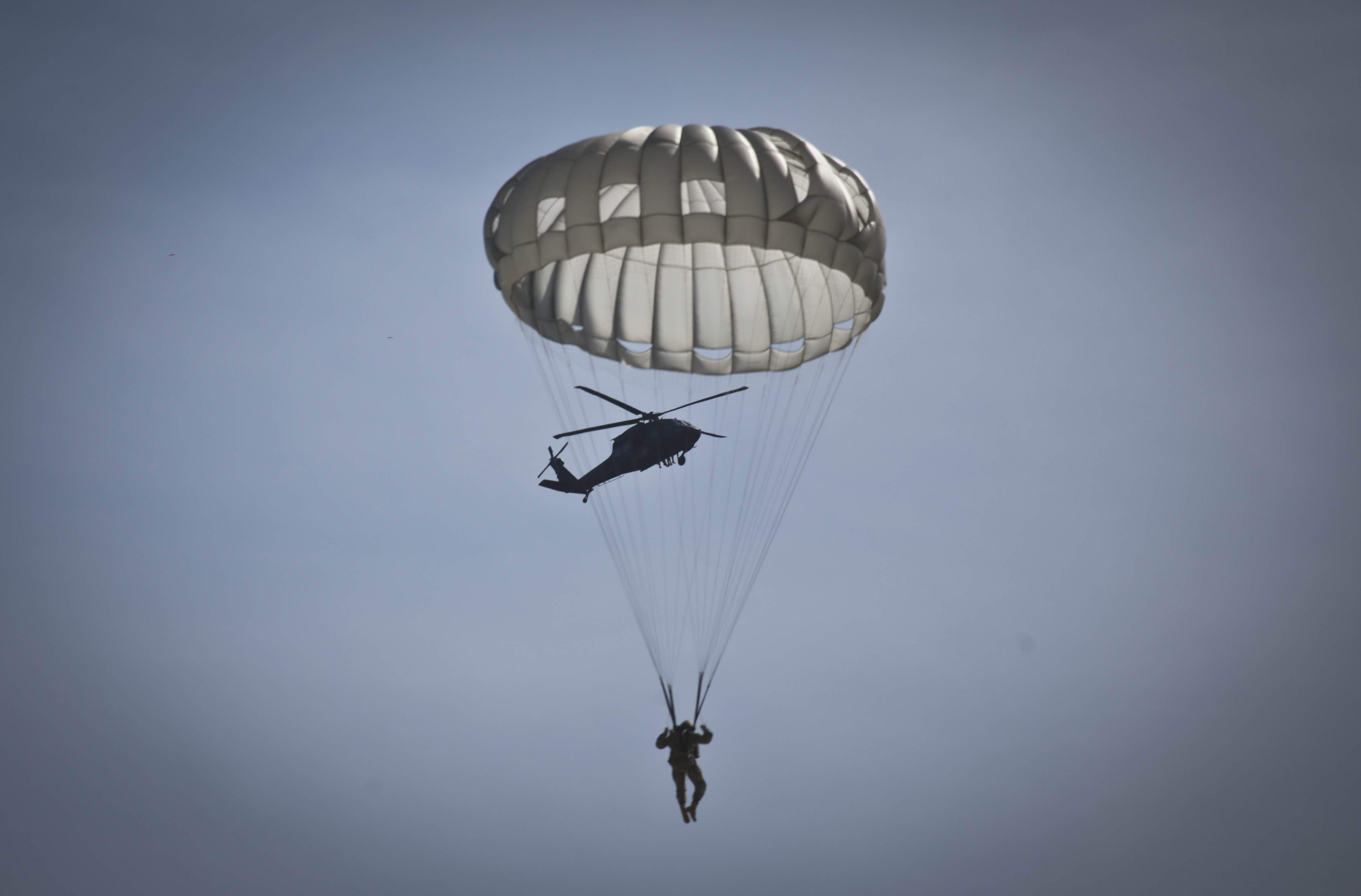 A U.S. Army Reserve paratrooper descends in his T-10 parachute as a New Jersey Army National Guard Black Hawk prepares to deploy more airborne Soldiers during a helicopter-borne parachute jump at Coyle Drop Zone, Joint Base McGuire-Dix-Lakehurst, N.J., March 12, 2016. (U.S. Air National Guard photo by Tech. Sgt. Matt Hecht/Released)