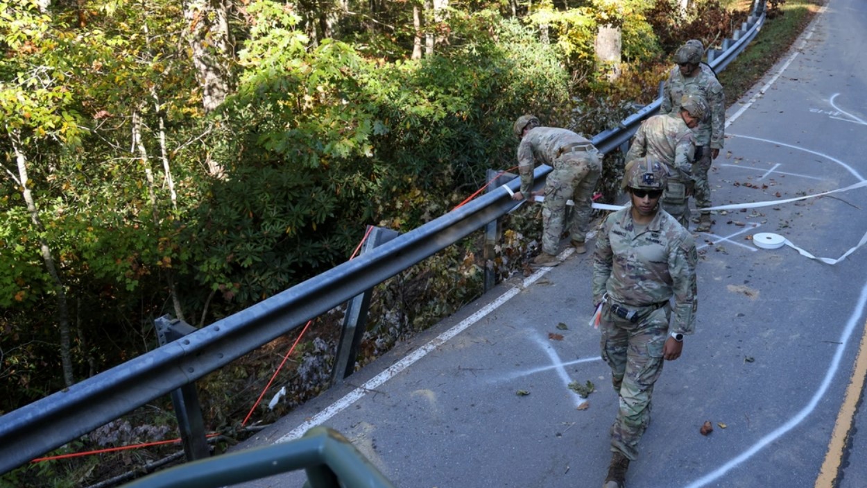 Soldiers walk along a damaged road, inspecting the barrier. The ground is marked to track their progress.