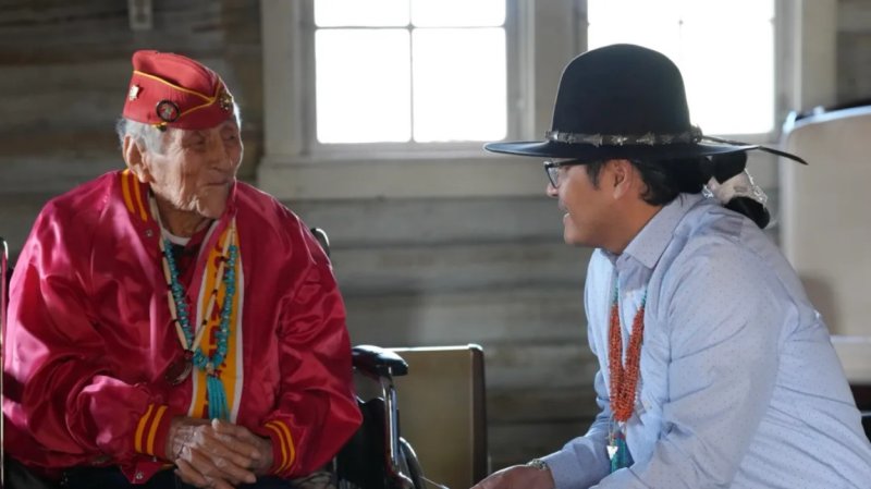 John Kinsel sits in a chair, wearing a red Navajo code talkers hat, next to someone wearing a wide brimmed hat.