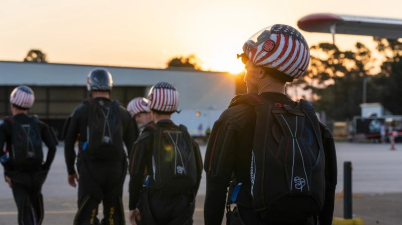 Four people in skydiving gear, wearing Americna flag-patterned helmets, walk towards a plane to skydive.