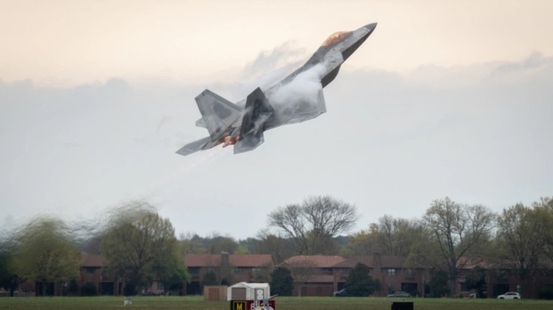 A F-22 fighter jet takes off a runway, air visibly disturbed by its speed.