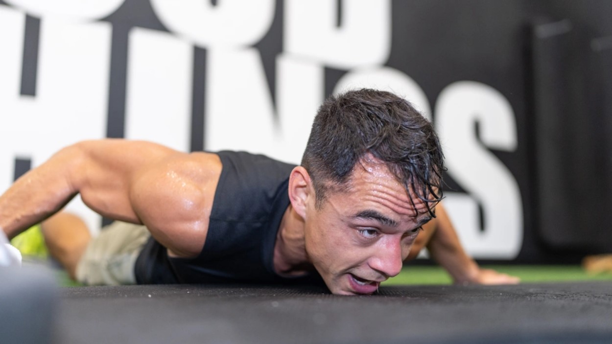 Tommy Vu lies on the ground at the bottom of burpee, chin on the ground, showing exhaustion and sweating.