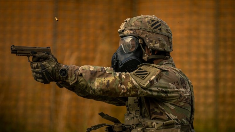 A soldier in a mask and goggles holds an M17 pistol with both hands.