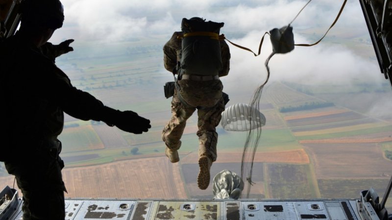 Troops jump from an airplane, parachutes streaming behind them.