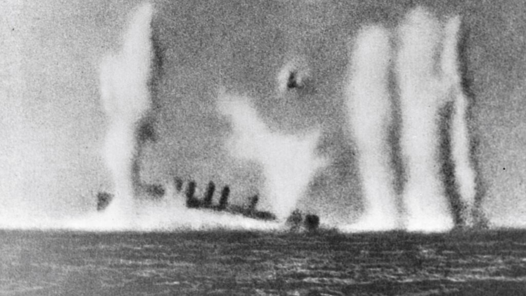 The USS Edsall sinking, smoke rising from the damage hull, while explosions kick up water around it.