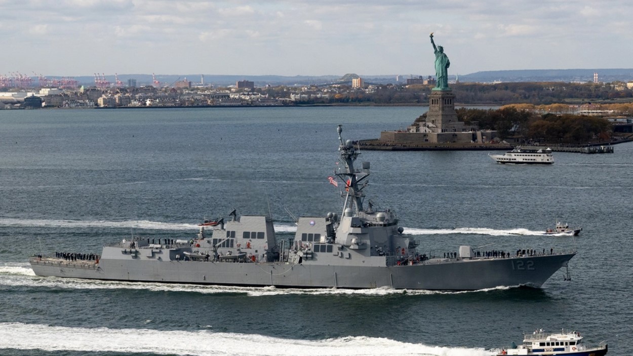 The destroyer the USS John Basilone sails through the waters around New York, wit the Statue of Liberty behind it.