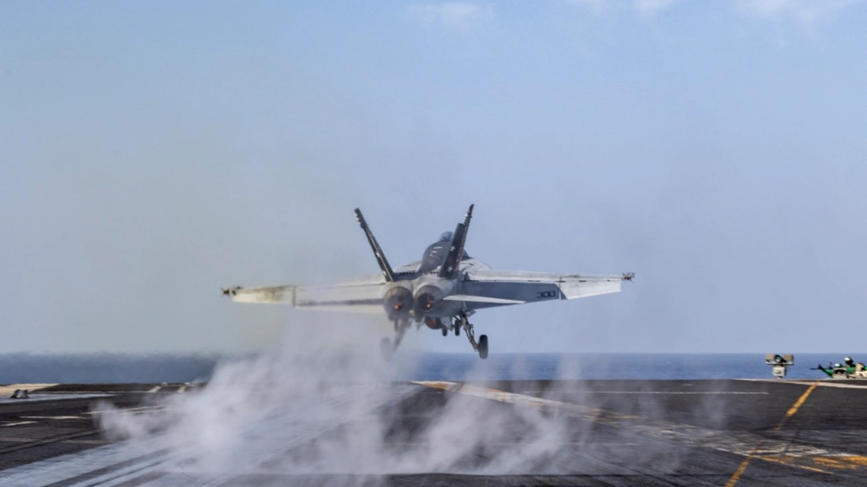 A F/A-18 fighter jet takes off from the deck of an aircraft carrier, smoke and steam trailing behind it.