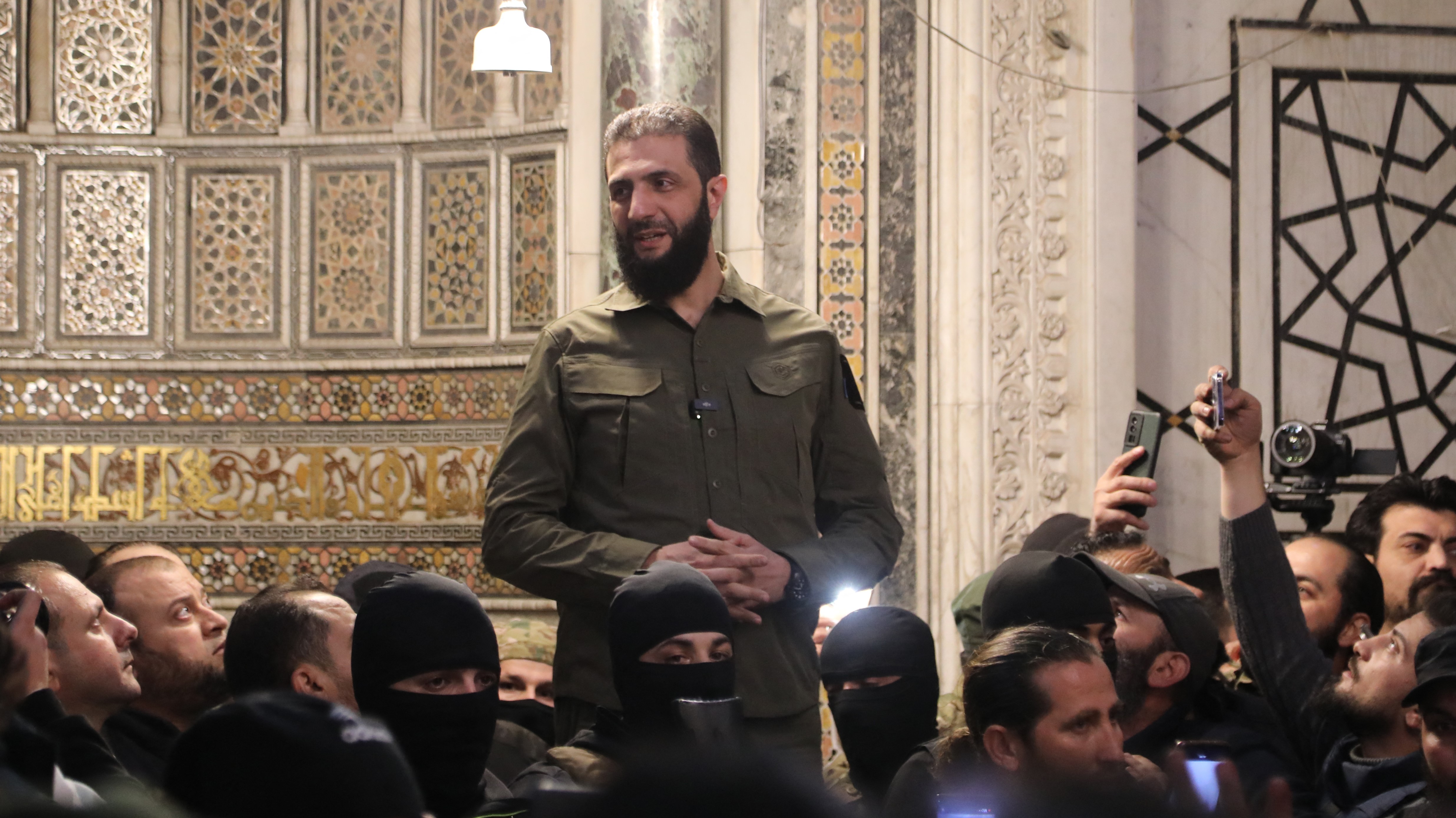 al-Julani, a bearded man in a military shirt, smiles and stands above supporters inside a mosque.