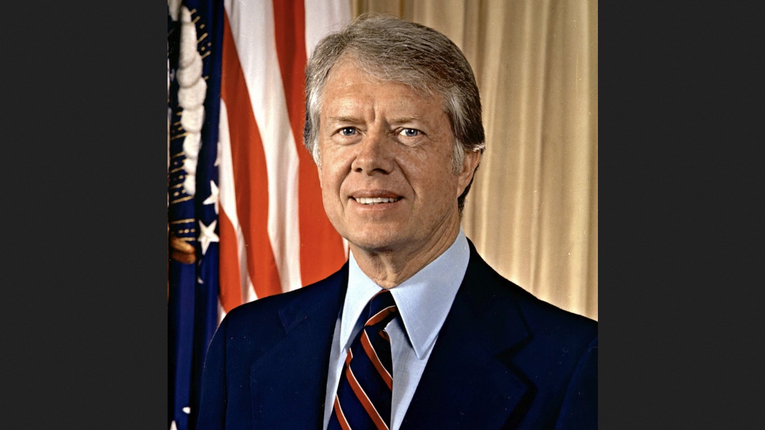 President Jimmy Carter, in a dark suit, with an American flag behind him