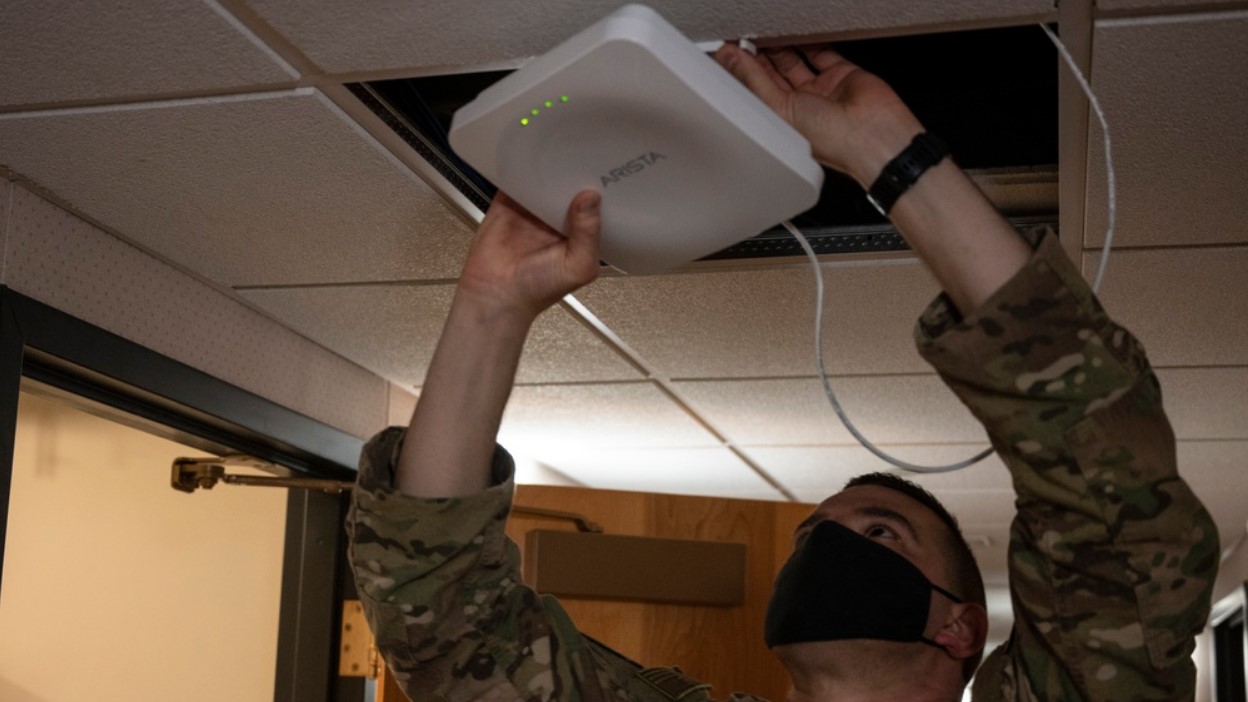 A man in military uniform with a face mask installs a white square Internet router in the ceiling of a barracks.