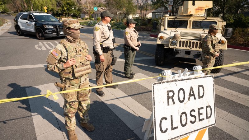 Soldiers in uniform alongside sheriff's deputies stand at a checkpoint with a "road closed" sign.