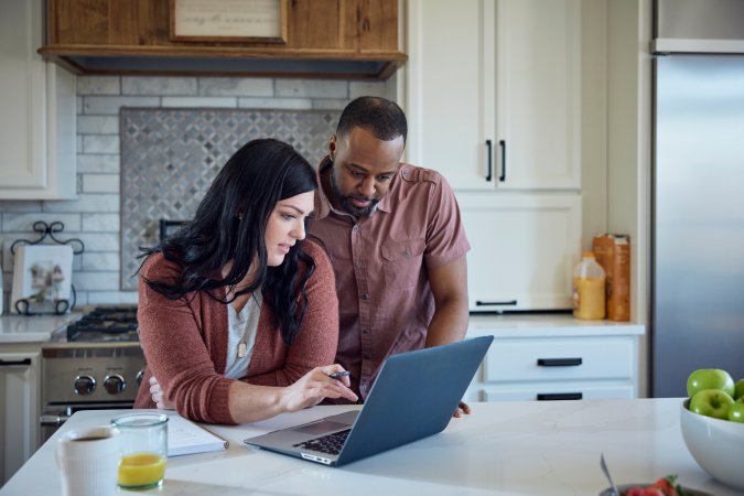 Caucasian woman and African American man sit at kitchen counter with breakfast working with pen, paper and laptop.