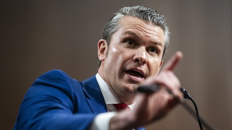 Pete Hegseth in a blue suit raises a finger next to his microphone.