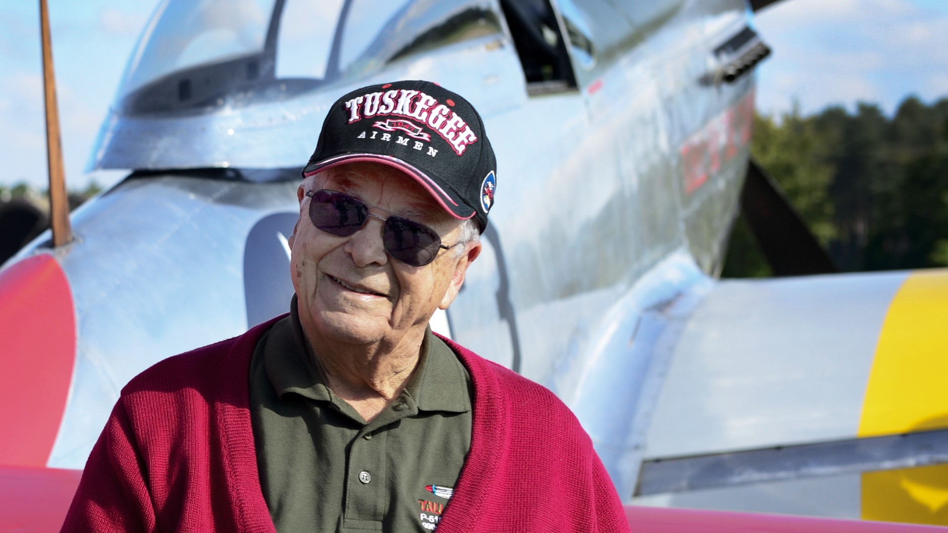 George Hardy, in a red sweater wearing a hat that says "Tuskegee," stands in front of his plane from World War II.