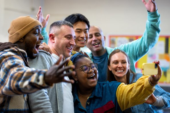 Waist-up shot of a group of friends taking a selfie together in a community centre. They are all smiling with positive emotion for the camera both wearing casual clothing. The community centre is located in Seaton Deleval in the North East of England.