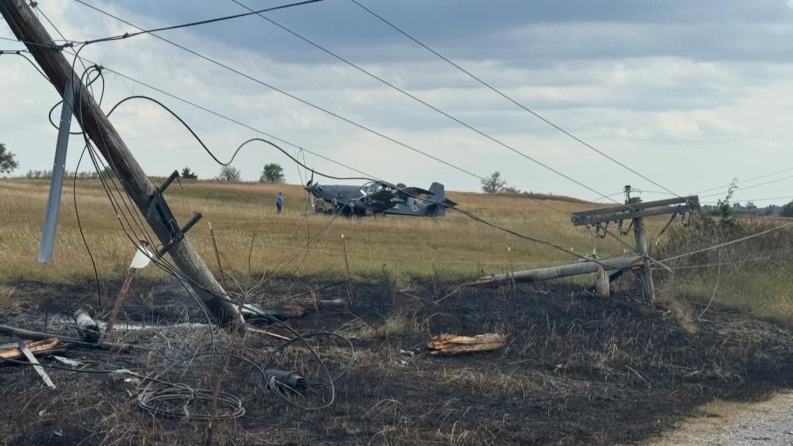 A turboprop plane rests in a field after crashing, two power poles askew in the front.