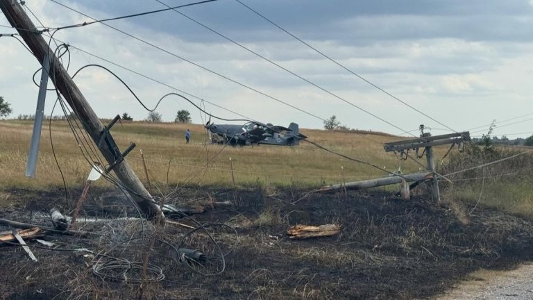 A turboprop plane rests in a field after crashing, two power poles askew in the front.