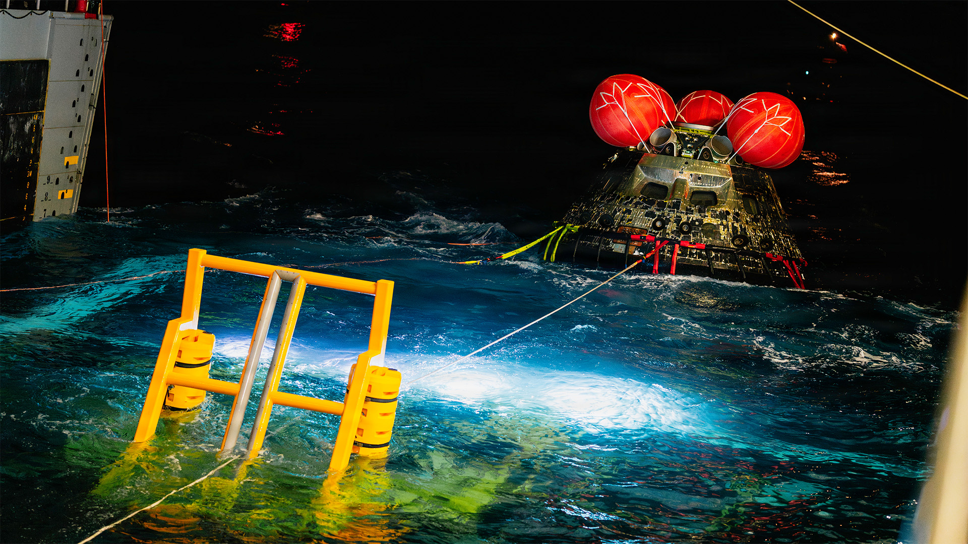 The NASA Orion space craft is winched into the well deck of the amphibious transport dock ship USS John P. Murtha, lit from below by NASA cameras inspecting the bottom of the capsule.