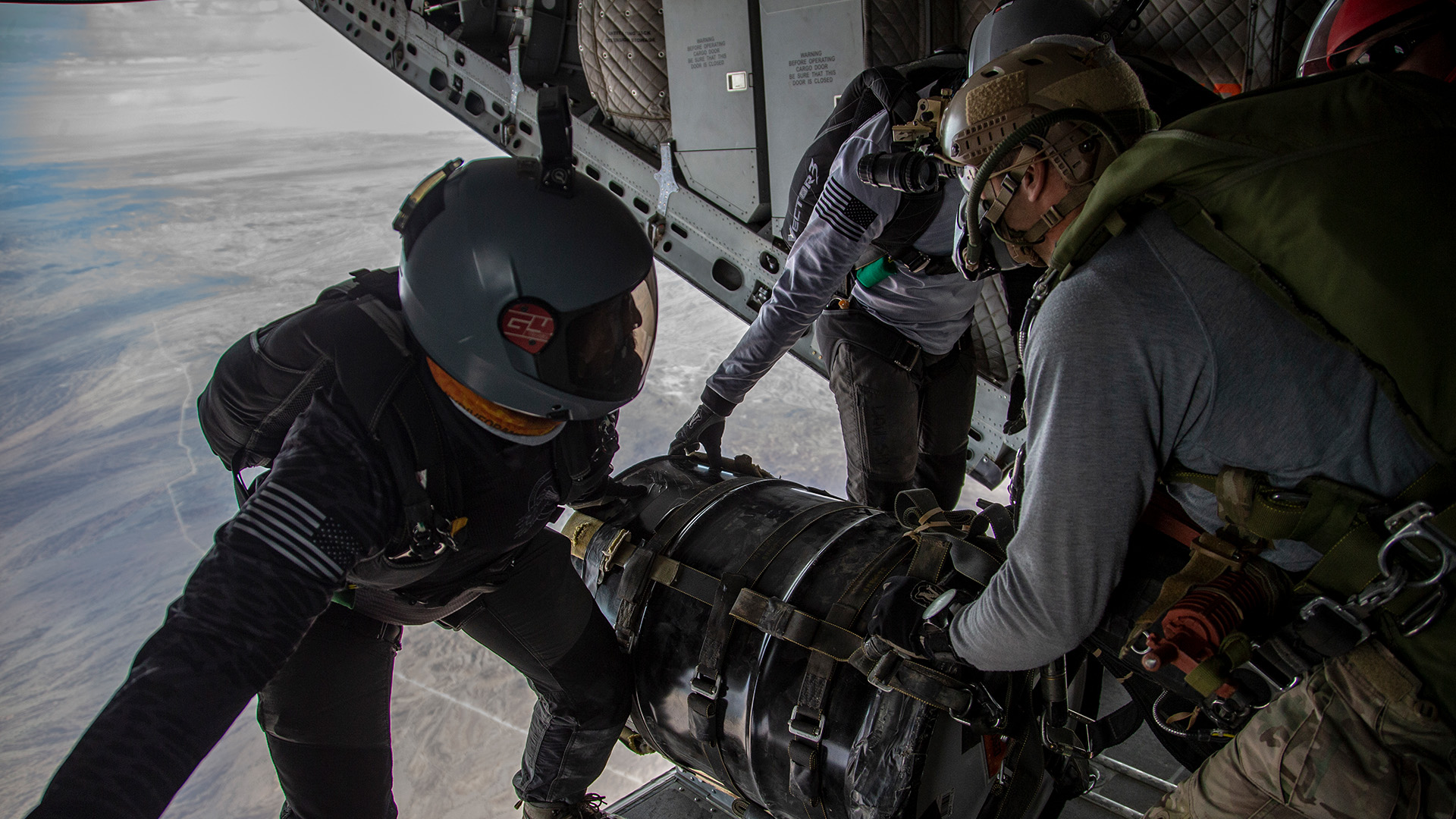 U.S. Marine and Army special operations troops prepare for a barrel jump, a tandem-style parachute operation in which a large, multi-hundred pound barrel hangs below a jumper as they fall.