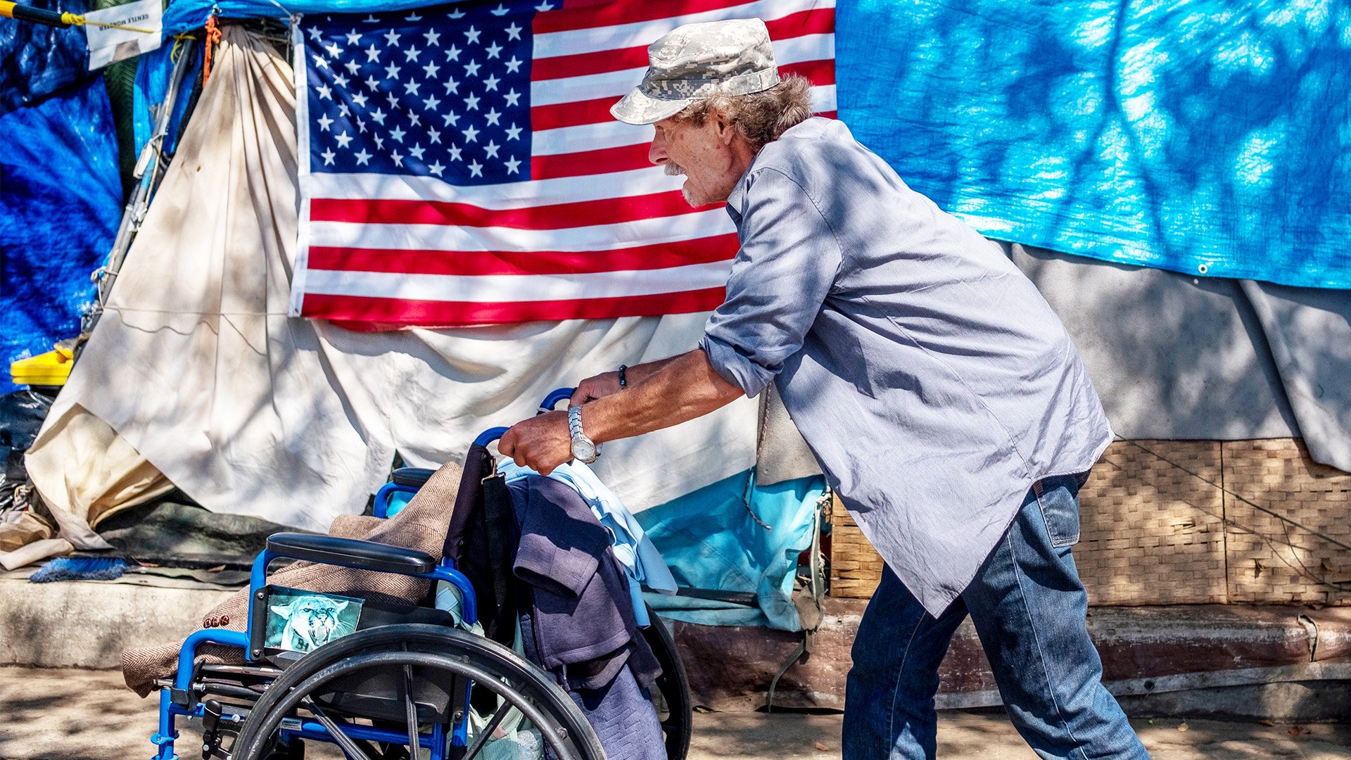 WEST LOS ANGELES, CA - AUGUST 30: Douglas Bue, 65, pushes his wheelchair to his tent next to a homeless encampment outside the West L.A. Veterans Affairs facilities on Monday, Aug. 30, 2021. The encampment is on San Vicente Boulevard in an unincorporated area near Brentwood in West Los Angeles, CA. Dozens of veterans live at the encampment, sometimes called Veterans Row, where some tents are decorated with U.S. flags. Recently a homeless veteran had been stabbed to death nearby. (Francine Orr / Los Angeles Times via Getty Images)
