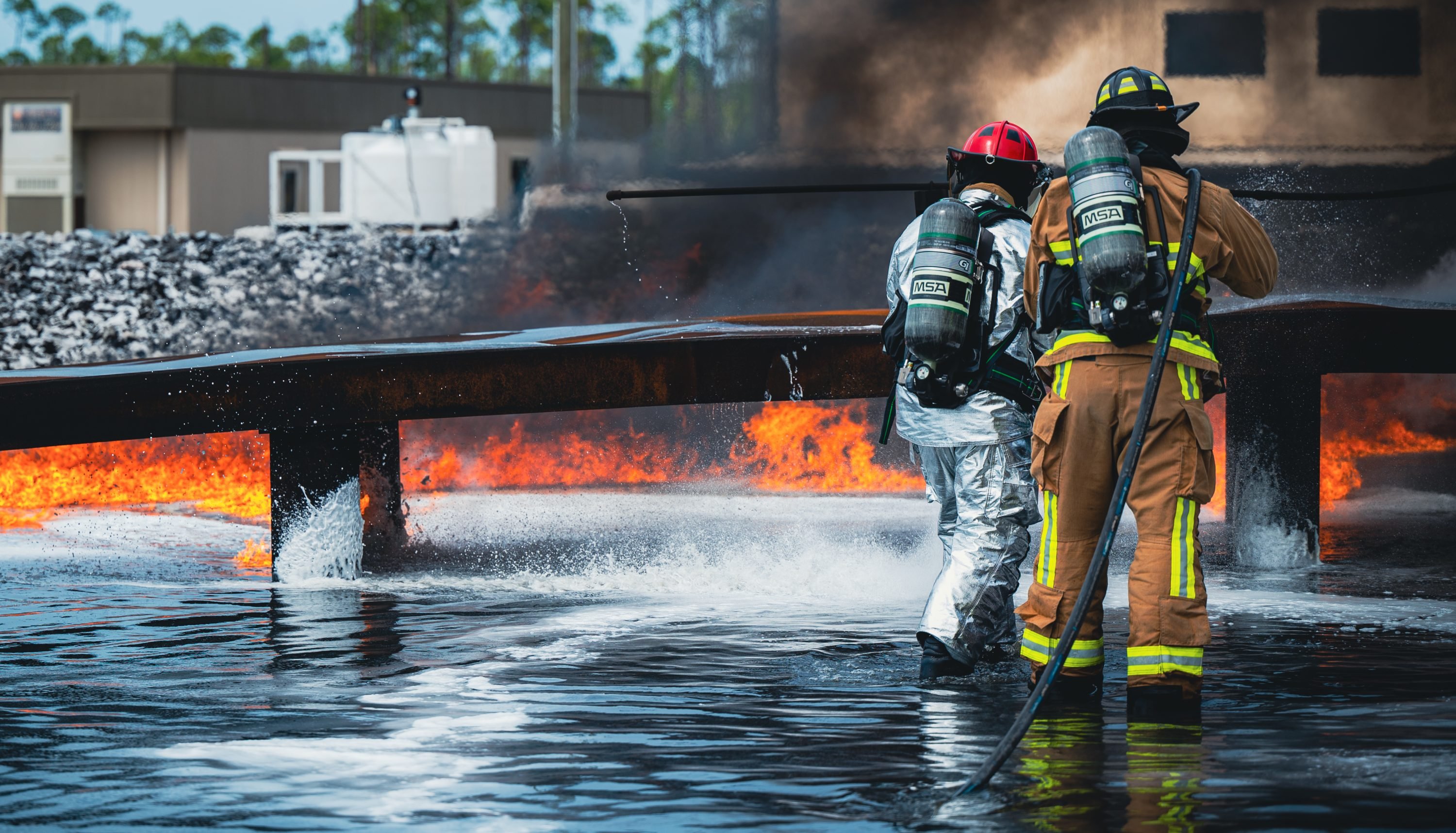 U.S. Air Force Staff Sgt. Christopher Collier, left, 801st Rapid Engineer Deployable Heavy Operational Repair Squadron Engineers Training Squadron fire protection cadre, and Airman 1st Class Jaremy Wright, 325th Civil Engineer Squadron fire protection firefighter, extinguish a controlled fire during a Fluorine-Free Foam training exercise within the Silver Flag burn pit at Tyndall Air Force Base, Florida, April 9, 2024. The chemical differences in the new foam caused significant changes in firefighting techniques with the transition. Rather than firefighters employing a “rainfall” technique to deploy foam and water through a hose at an arching angle, they now shoot directly at the base of the fire. (U.S. Air Force photo by Airman 1st Class Zeeshan Naeem)